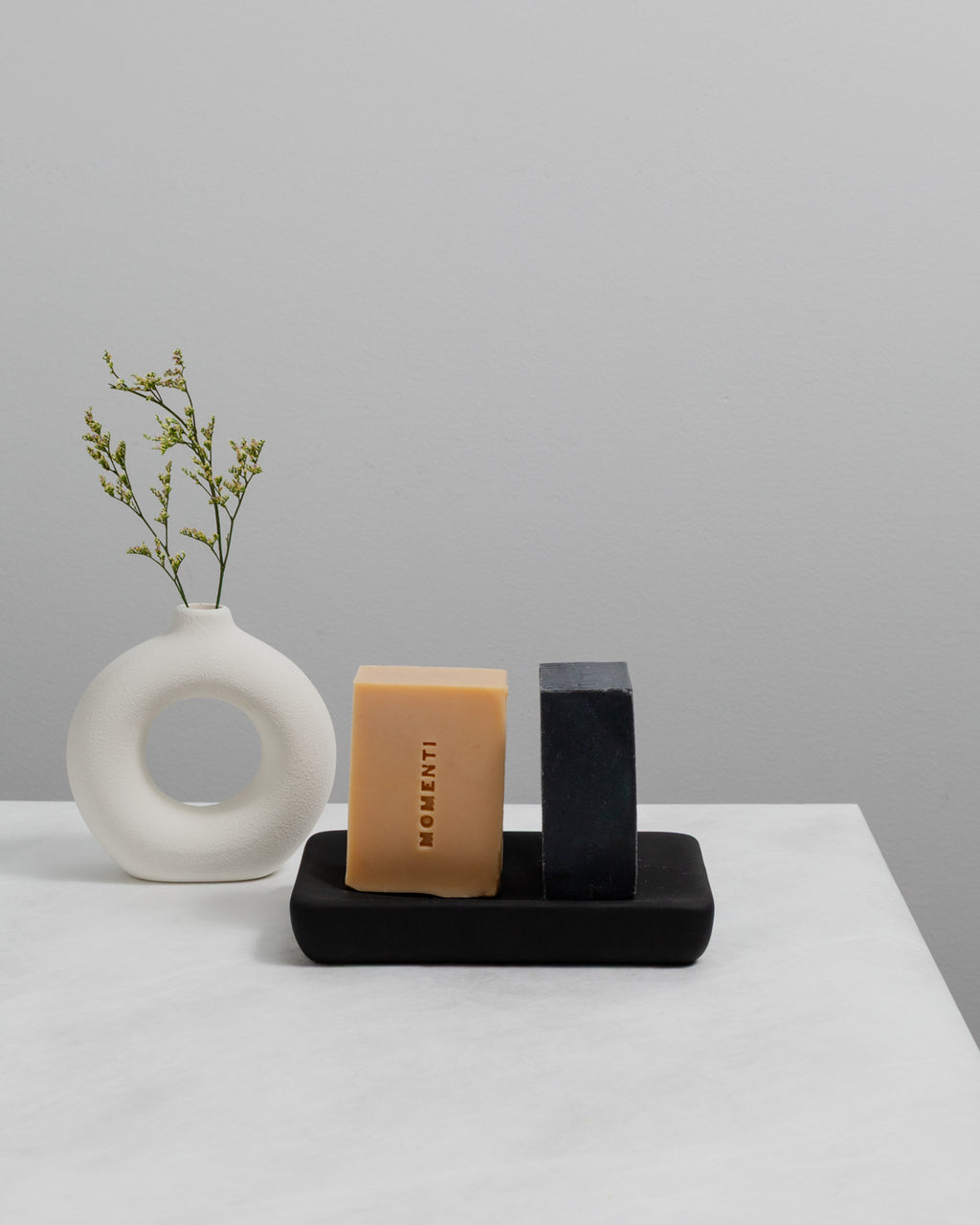 White vase with greenery, orange bar of soap, and black block on a white surface with gray background