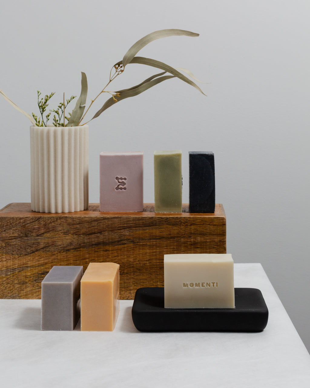 Collection of soap bars on a wooden shelf with a vase and plant in the background.