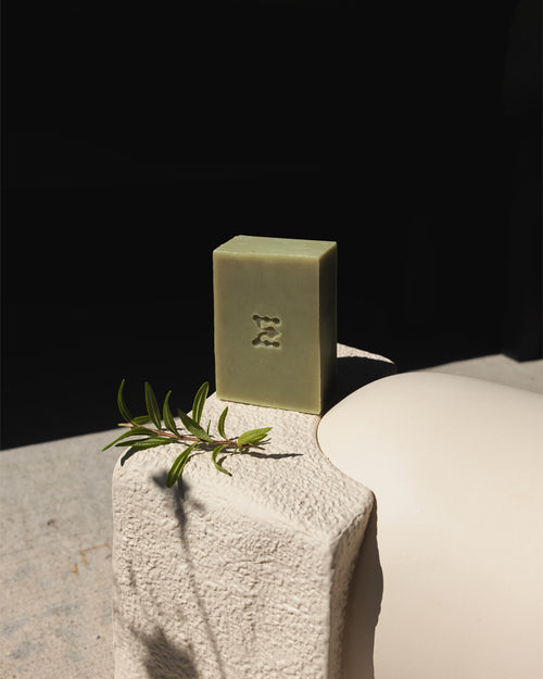 Natural green rosemary soap bar standing against a dark background standing on a white concrete surface with natural fresh rosemary stem next to it. 