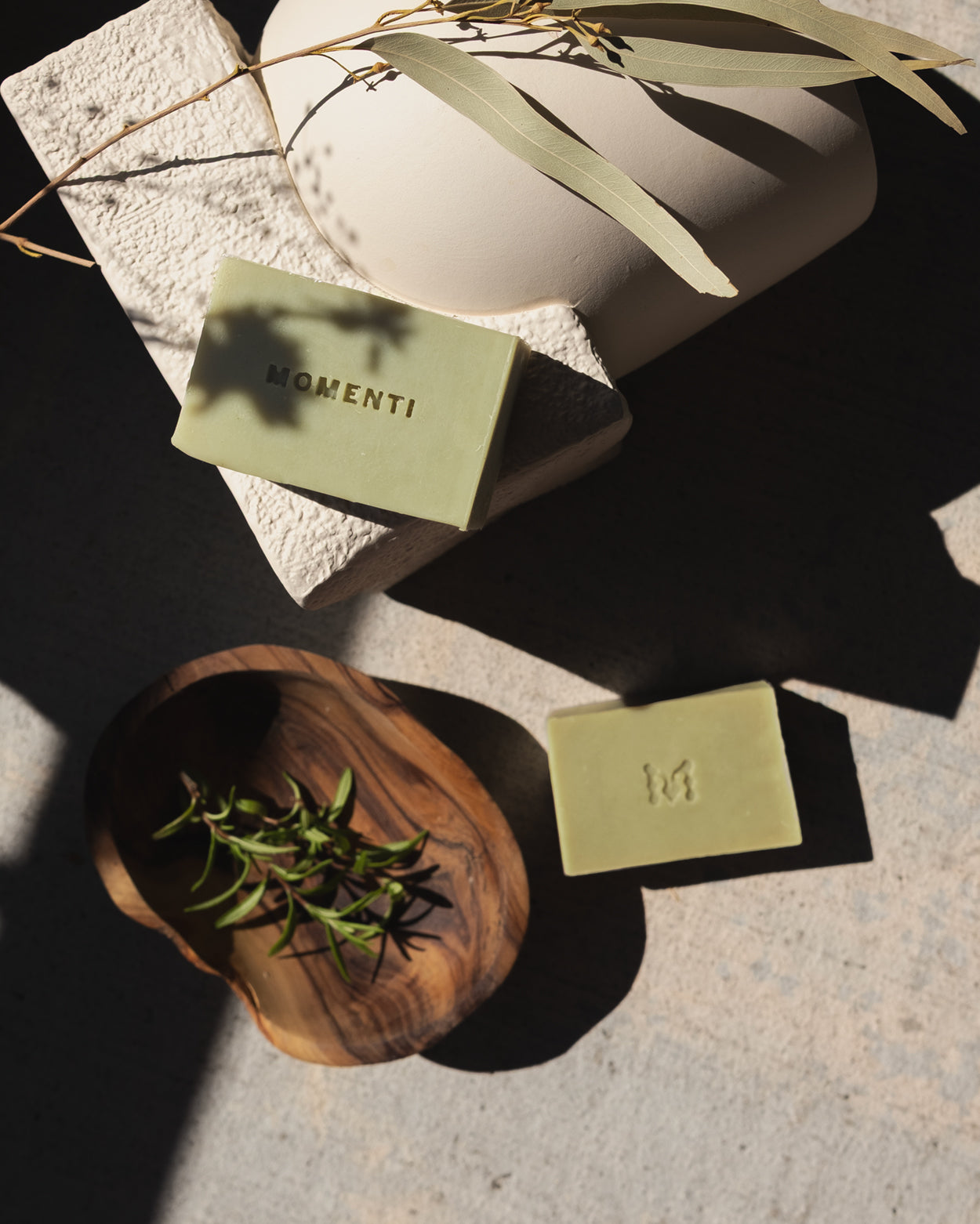 Two green rosemary natural soaps photographed outside in sunlight with stems of fresh rosemary in a natural wood bowl. 