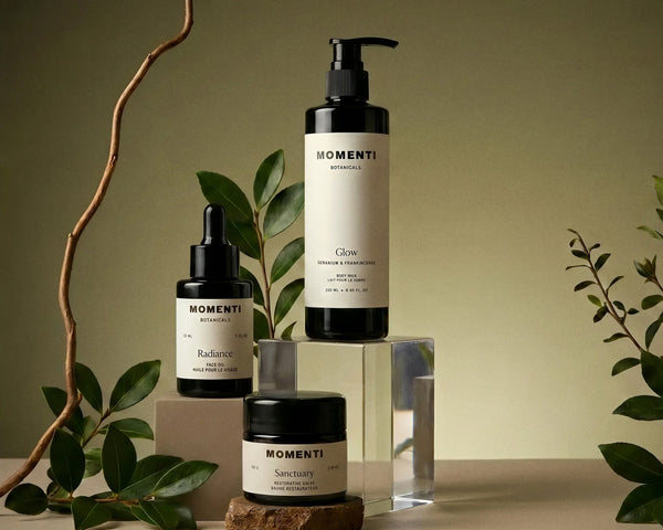 Cosmetic skincare products such as face oil, body lotion, and skin balm with 'Momenti' labels on a neutral background with plants.