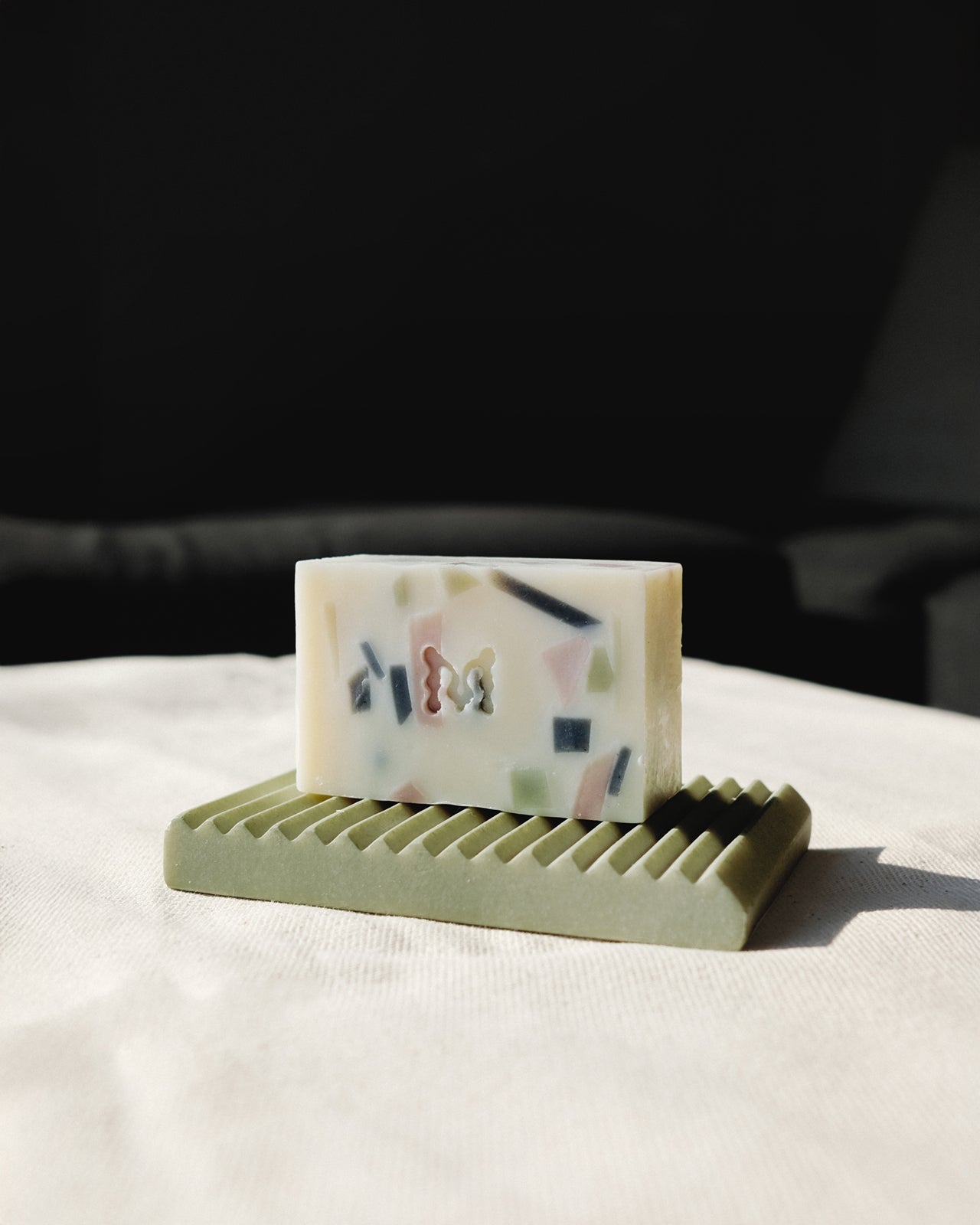 Natural speckled Costiera soap bar on a green soap dish sitting on a beige canvas cloth